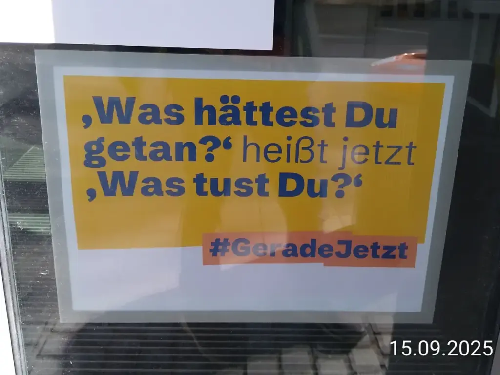 Ein Foto aufgenommen mit einem Smartphone. Ein DIN A4 Blatt bedruckt an einer Fensterscheibe. Das Blatt ist weiß. Der Hintergrund mit dem Text in blauer Farbe "Was hättest Du getan?" heißt jetzt "Was tust du?" ist orange.Zwischen dem Hintergrund in Orange und Weiß steht #GeradeJetzt Rechts unten ist der digitale Zeitstempel 15.09.2025