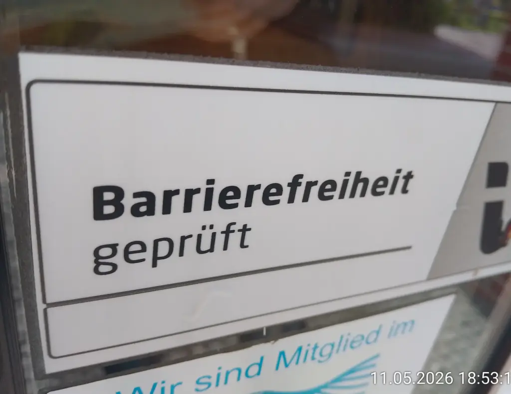 Foto von einem Aufkleber an einer Glastür. Auf diesem Aufkleber steht"Barrierefreiheit (fett geschrieben) geprüft" vor einem weißen Hintergrund. Rechts daneben ist der Hintergrund grau / abgedunkelt und es ist Ansatzweiße ein "Haken" / "Check" in Schwarz und den Farben rot und gelb zu sehen". Darunter ist ein weitere Aufkleber, auf welchem steht "Wir sind Mitglied im" und darunter ist ein Symbol, welches nichts ganz ersichtlich ist zu sehen. Rechts unten im Fotos steht 11.05.2026 18:53