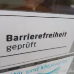 Foto von einem Aufkleber an einer Glastür. Auf diesem Aufkleber steht"Barrierefreiheit (fett geschrieben) geprüft" vor einem weißen Hintergrund. Rechts daneben ist der Hintergrund grau / abgedunkelt und es ist Ansatzweiße ein "Haken" / "Check" in Schwarz und den Farben rot und gelb zu sehen". Darunter ist ein weitere Aufkleber, auf welchem steht "Wir sind Mitglied im" und darunter ist ein Symbol, welches nichts ganz ersichtlich ist zu sehen. Rechts unten im Fotos steht 11.05.2026 18:53