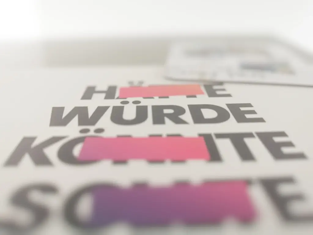 Foto einer weißen Postkarte an einer Kühlschranktür mit Perspektive von unten. Auf der Postkarte stehen folgende Wörter: Hätte (durchgestrichen) Würde, Könnte (durchgestrichen) Sollte (durchgestrichen) untereinander. Hätte steht ganz oben und Sollte steht ganz unten. Das Wort Würde ist am besten lesbar, alles andere ist verschwommen.