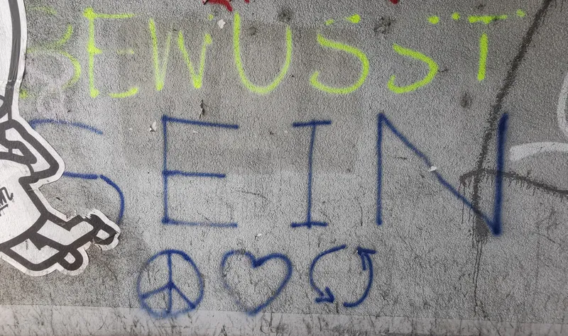 Mauer / Wand mit dem Hand geschriebenen Graffiti in gelber Farbe "Bewusst" und darunter in blauer Farbe in gleicher Breite "Sein". Darunter 3 Symbole: Das Peace-Zeichen / Friedenssymbol, Herz, und Recycling / Refresh / Ganzheitlich