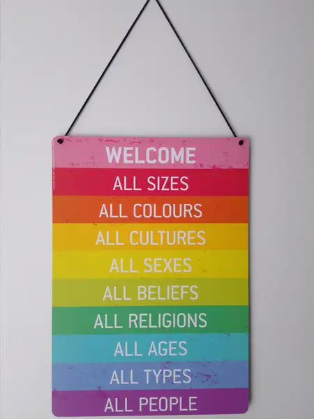 Ein "Schild" hängt durch eine Schnur an einer weißen Wand. Ganz oben steht das Wort "Welcome" in weißer Schrift vor farbigen Hintergrund. Darunter stehen jeweils unterschiedliche Bereiche - alle Beginnend mit "All" auch in weißer Schrift mit jeweils anderer Hintergrundfarbe. Sizes Colours Cultures Sexes Beliefs Religions Ages Types People