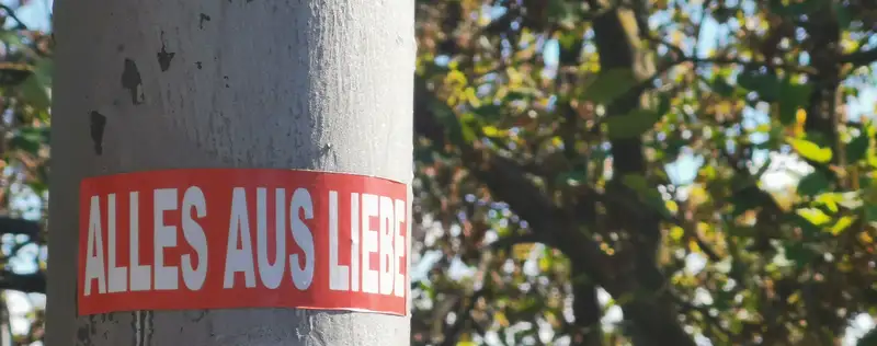 Ein Aufkleber mit rotem Hintergrund und weißen Buchstaben "Alles aus Liebe" in Groß Buchstaben an einem grauen Laternen Oberfläche. Wird von rechts von der Sonne erhellt. Im Hintergrund ist unscharf ein Baum mit grünen Blättern und ein blauer Himmel zu sehen.