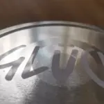 Ein grauer / silberner Deckel mit dunklen & braunen Hintergrund. Linke eite vom Deckel ist heller und zur rechten Seite wird es dunkler. Darauf steht in weißer Schrift "Glück".