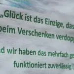 Foto von einem Stück Papier in weißer Farbe mit grünem Text. Ein leichter Unschärfeverlauf von Links scharf, nach rechts unschärfer. Darauf steht "Glück ist das Einzige, das sich beim Verschenken verdoppelt". (Und wir haben das mehrfach getestet - funktioniert zuverlässig!)