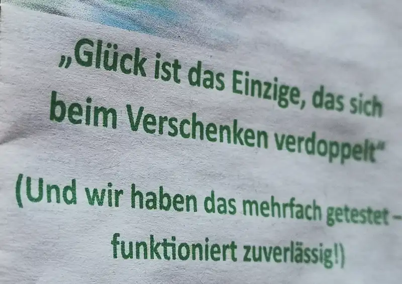 Foto von einem Stück Papier in weißer Farbe mit grünem Text. Ein leichter Unschärfeverlauf von Links scharf, nach rechts unschärfer. Darauf steht "Glück ist das Einzige, das sich beim Verschenken verdoppelt". (Und wir haben das mehrfach getestet - funktioniert zuverlässig!)