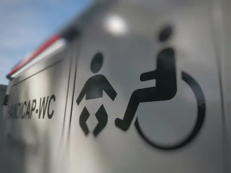 Tagsüber und blauer Himmel. Ein Container steht draußen mit dem schwarzen Symbol für Rollstuhl und Baby auf hellen Hintergrund an der Wand. An der Tür steht "Handicap-WC". Über diesem Wort ist eine leichte Beleuchtung installiert