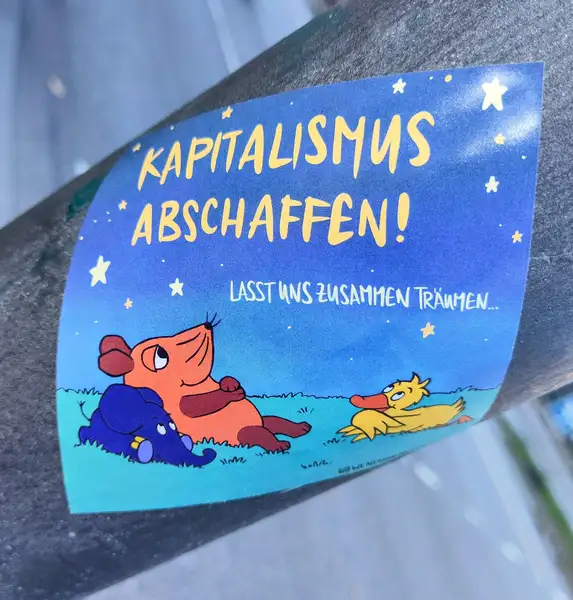 Ein Aufkleber ist auf einer Metalisch aussehenden Oberfläche zu sehen welche Rund ist. Der Hintergrund der Metalischen Gegenstand in Unscharf und ist nicht genau erkennbar. Auf dem Aufkleber ist eine Comic-Artige Umgebung zu erkennen in der Natur. Im Sternenhimmel steht "Kapitalismus abschaffen! in gelber Schrift. Darunter steht in weißer Farbe Lasst und zusammen träumen... Eine Maus, ein Elefant und eine Ente liegen entspannt auf einer Wiese und schauen zu diesen Worten